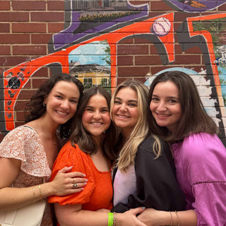 Four women posing together in front of a colorful mural on a brick wall.