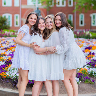 Four women in white dresses standing together in front of a brick building and flower garden.