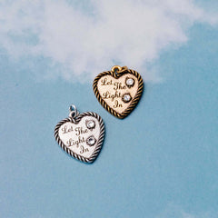 &LIVY Let The Light In Heart Charm in both finely finished gold and finely finished silver featuring heart-shaped pendants with Let The Light In engraving Austrian crystal accents and rope edge detail on blue sky background