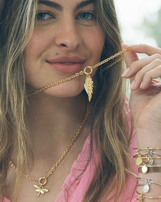 Woman wearing a gold necklace with an angel wing pendant, surrounded by more gold jewelry.